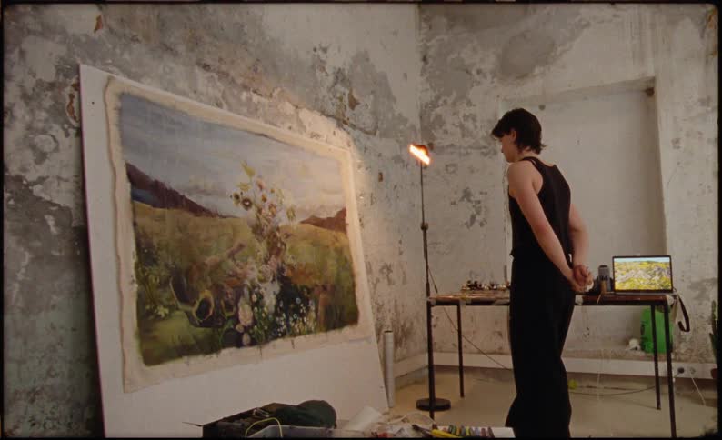 A man wearing a black singlet and black trousers stands in an art studio staring at a large painting resting against the wall as a light beside a table illuminates the artwork with a laptop also placed on the table