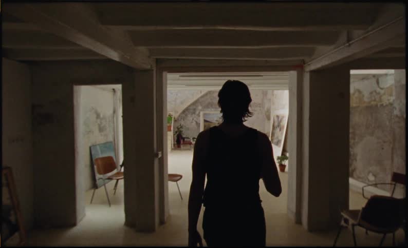 Woman with a pot walking down the stairs towards a room with art paintings and interacting with a laptop