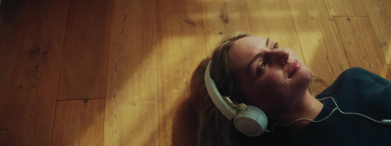 A young blonde woman lies on a wooden floor wearing wired headphones smiling contentedly
