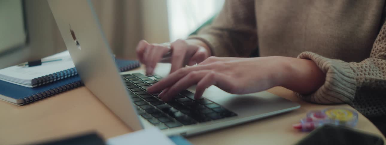 A woman types on a laptop at a desk with notebooks a smartphone and correction tape scattered nearby