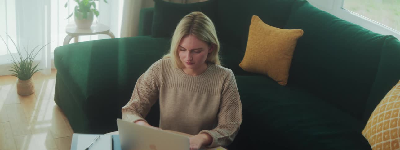 A young blonde woman wearing a sweatshirt is seated on a couch typing on the keyboard of a laptop placed on the table in front of her at midday