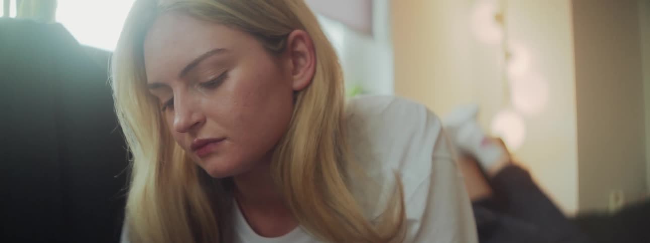 A young blonde girl with long hair lies on a couch looking down while gently twisting her head from left to right