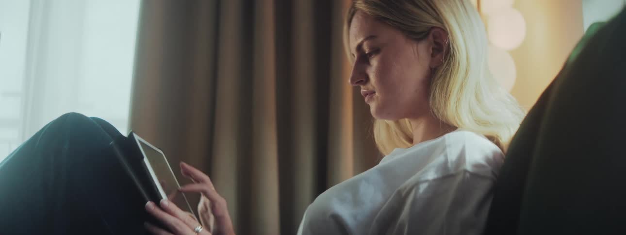 A woman with blonde hair is seated comfortably holding and focusing on a tablet with beige curtains and soft lighting in the background