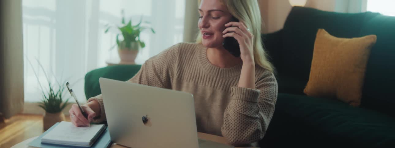 A blonde woman sits in her living room while talking on a smartphone