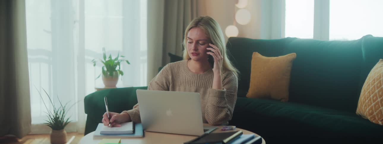 A blonde woman writes in a notebook while talking on her smartphone