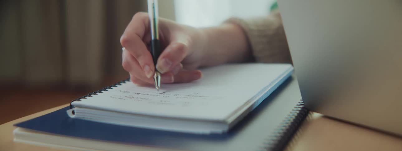 A hand writes in a notebook beside an open laptop with visible text on the notebooks pages