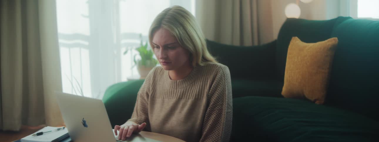 A blonde woman sits on her living room floor and scrolls on a laptop