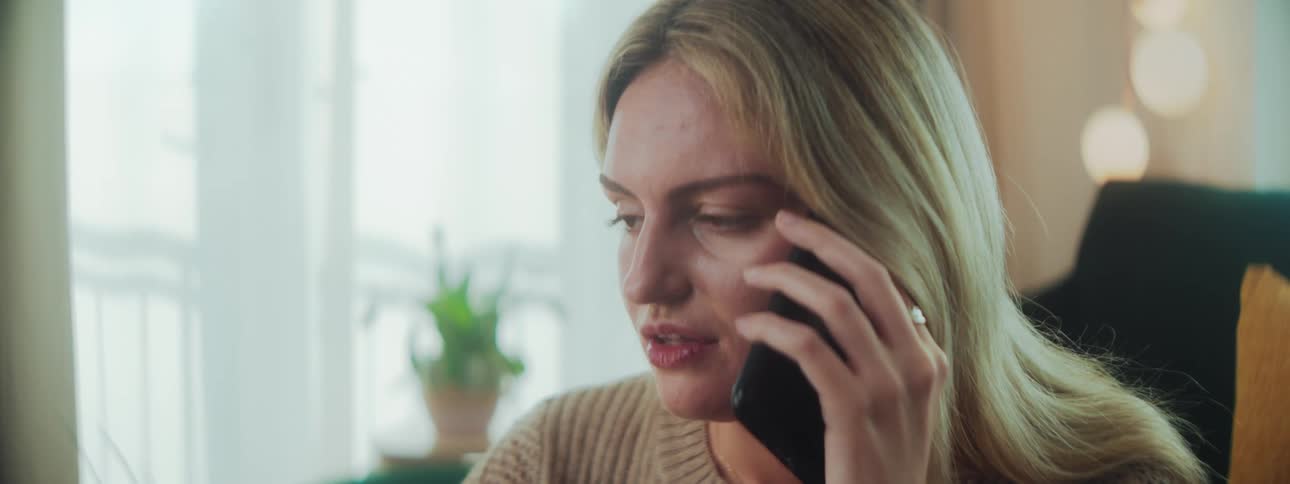 A woman with long blonde hair holds a phone to their ear wearing a beige sweater with a potted plant and warm ambient lighting in the background