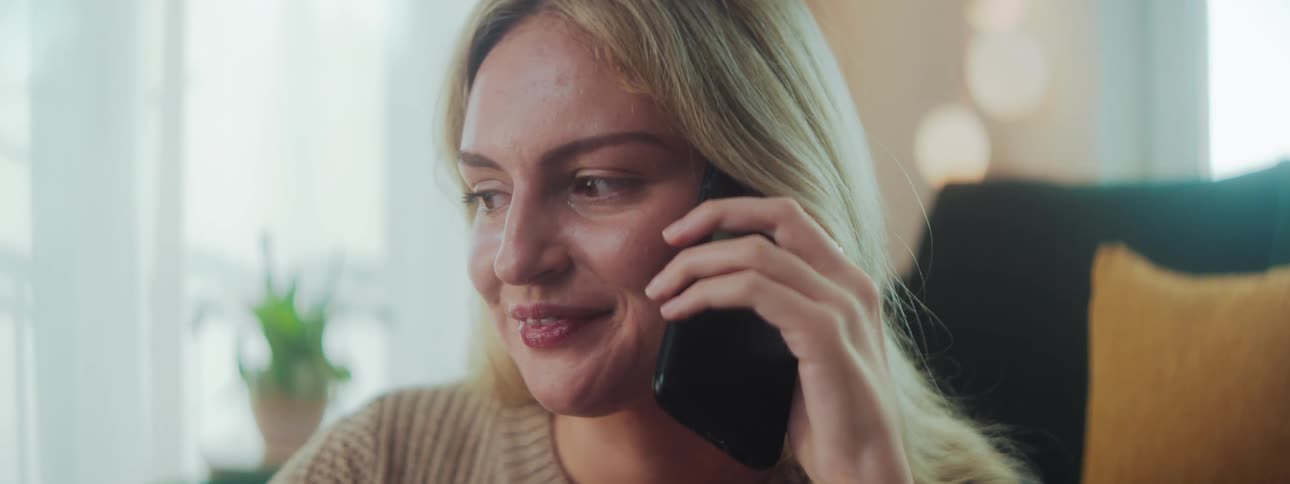 A woman wearing a beige sweater holds a phone to their ear with indoor plants and soft light in the background