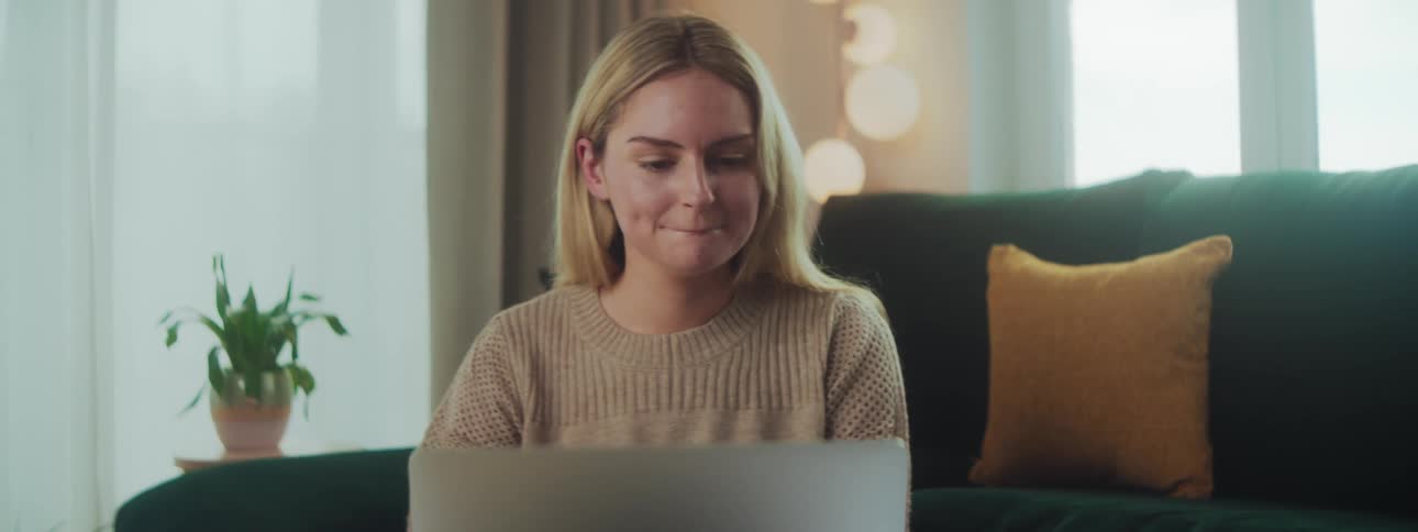 A woman on a video call seated on a green couch with a yellow pillow a potted plant and warm wall lights in the background