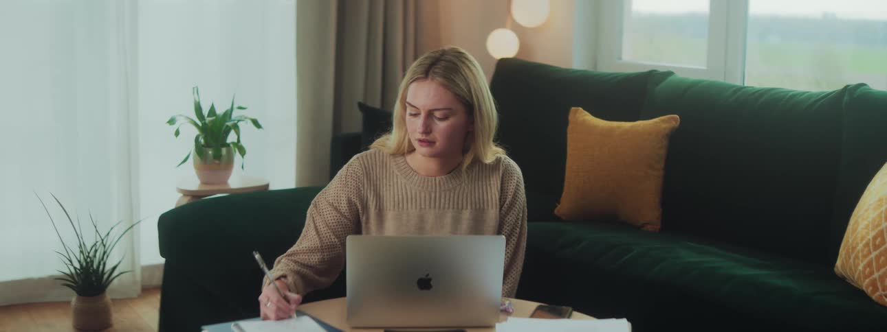A woman in a room at a table writing in a notebook while looking at her laptop