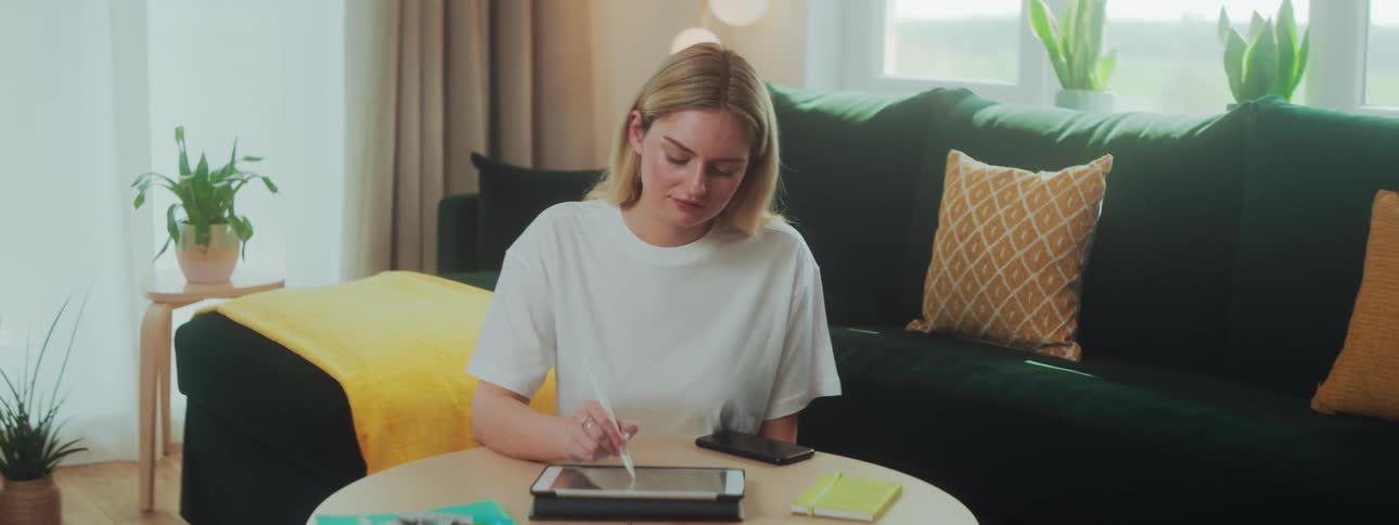 A woman in a room sitting at a table writing on her tablet with a stylus