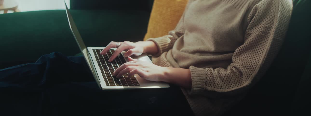 A woman is sitting on a couch typing on a laptop keyboard at midday