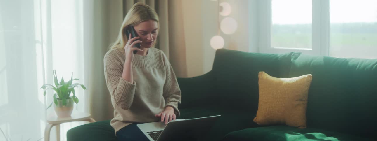 A woman in a room with a laptop on her lap picking up a phone to talk