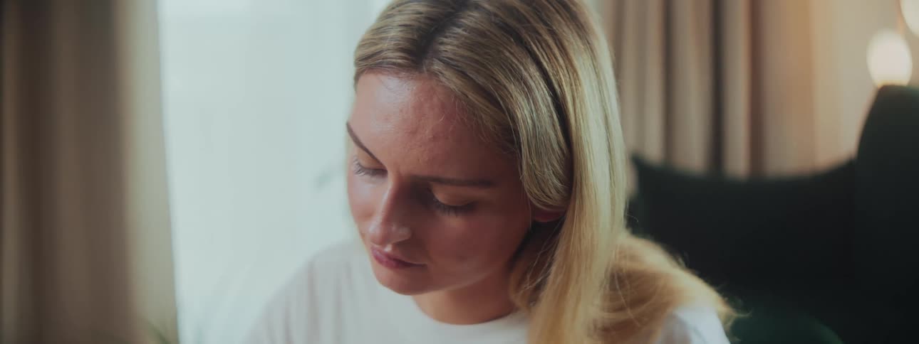 A young blonde woman sits quietly glancing down as she slowly turns her head from left to right