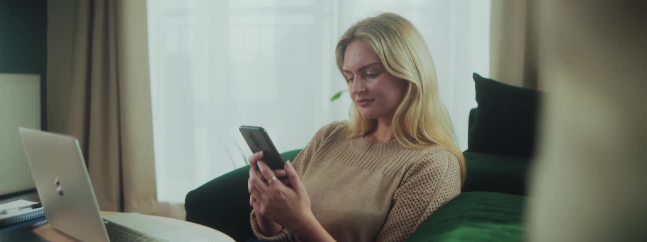 A woman is sitting on a couch at midday browsing her smartphone with a laptop placed on the table in front of her