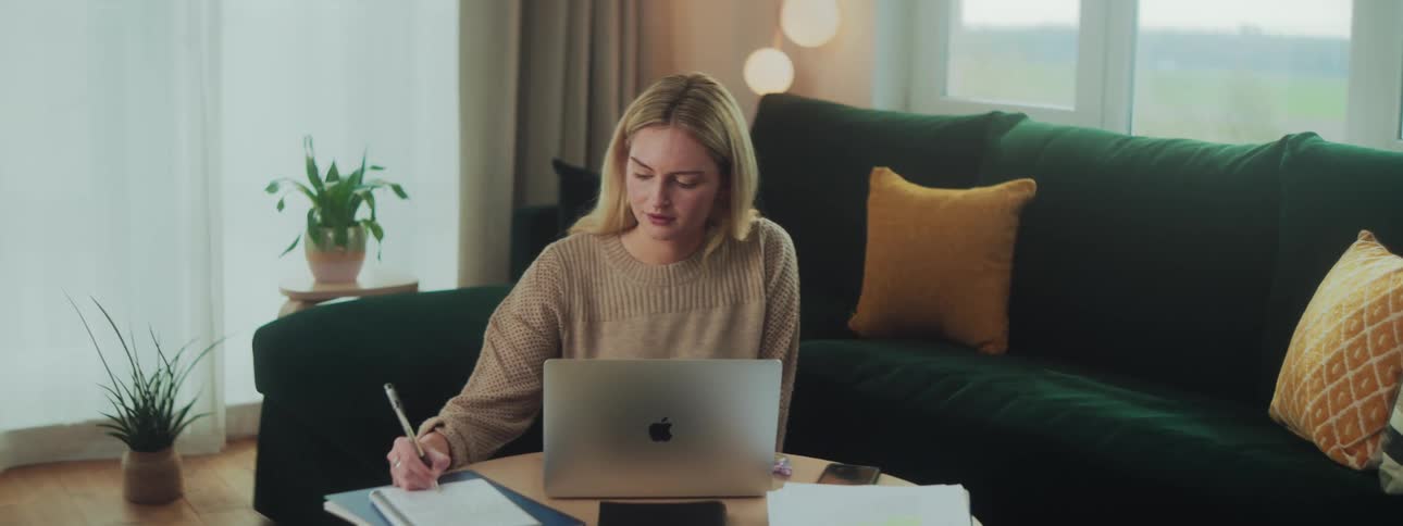 A blonde woman sits on the floor writing in a notebook with a pen while studying with her laptop placed on a table in front of her in the living room at midday