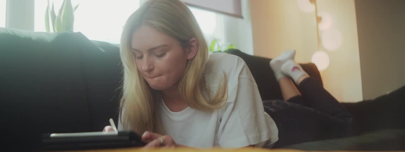 Blonde woman lying on the couch face down drawing on a tablet