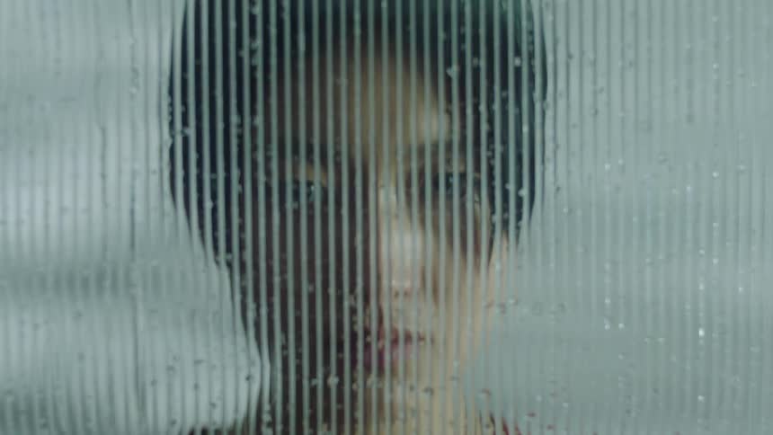 A person stands behind a glass panel streaked with vertical lines and dotted with water droplets distorting their features