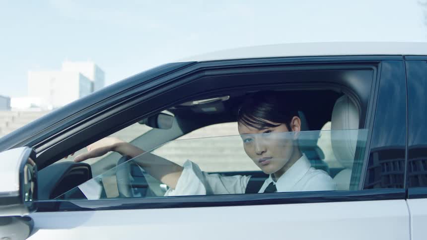 An asian woman sits in the drivers seat of a parked car beside the road at midday wearing a white collared shirt as she winds down the window