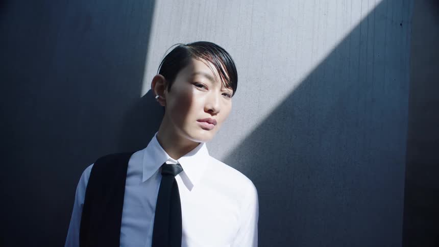 A person with a sleek short haircut and an earring dressed in formal attire stands near a wall casting a distinct sharp shadow