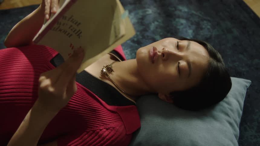 A young woman with dark hair wearing a red top and a necklace lies on a gray pillow while reading a book