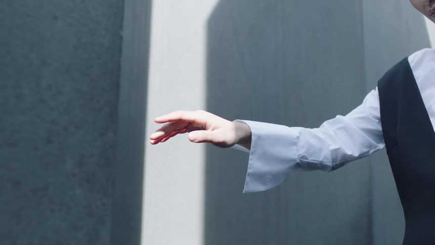 A person in a white shirt and dark vest extends an arm with an open hand in front of a concrete wall with shadows
