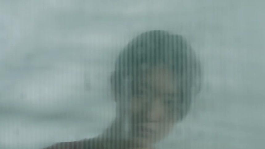 An asian man is seen through a blurred glass with water dripping down its surface as he moves his head seductively