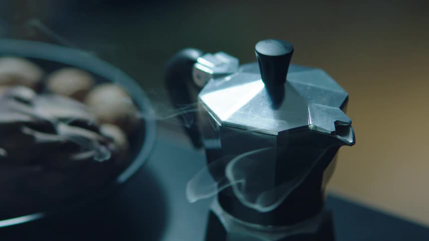 A stovetop espresso maker releases steam beside a bowl filled with walnuts