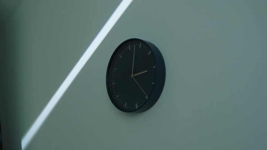 A black circular clock with gold hands hangs on a wall as a beam of sunlight moves across it