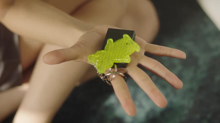 A close up of a hand holding a yellow glossy cartoon bear shaped keychain attached to a black box possibly a controller via a silver keyring and chain