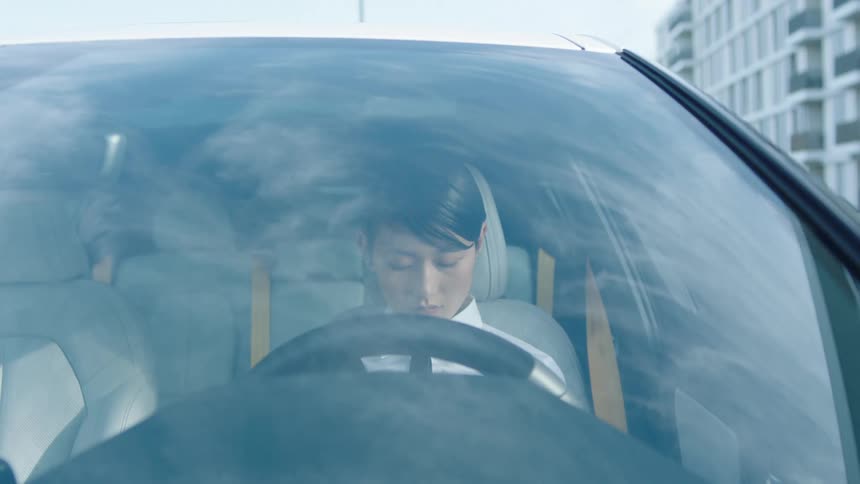 An asian young woman wearing a white collared shirt sits in the drivers seat of a car looking sad