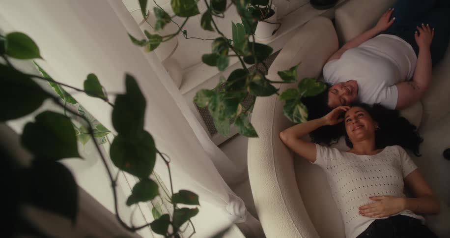 Two women lying on a couch one gently caressing the other as they both smile in a romantic moment