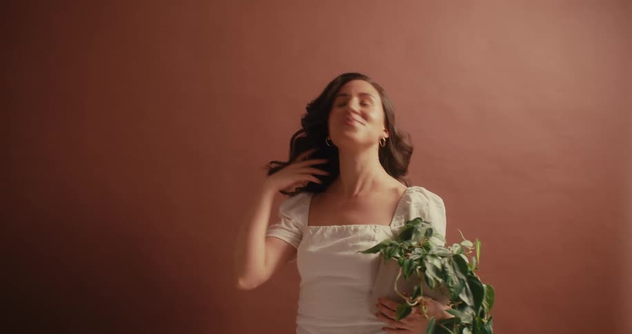 A woman dressed in a white blouse holds a vibrant green potted plant against a warm earthy backdrop