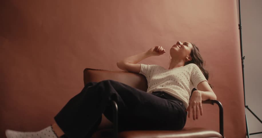 A woman sits in a chair with her legs hanging off the arm of the chair while running her hand through her hair