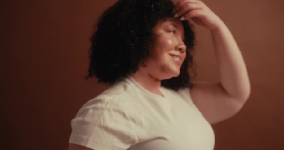 A chubby woman with curly hair wearing a white tshirt and a tattoo on her right arm laughs joyfully while playing with her hair