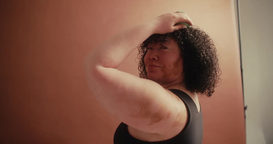 A person with visible skin variations holds a green object to their curly hair while looking at the camera
