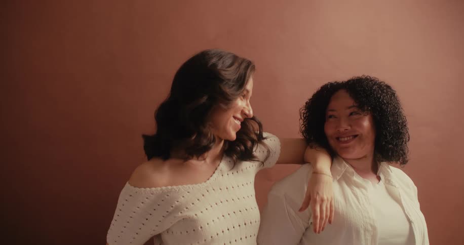 Two women posing against a wall exchange smiles briefly