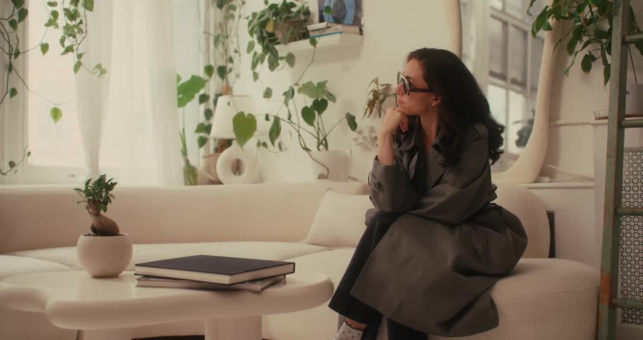 A lady sitting on a couch in a trench coat and sunglasses