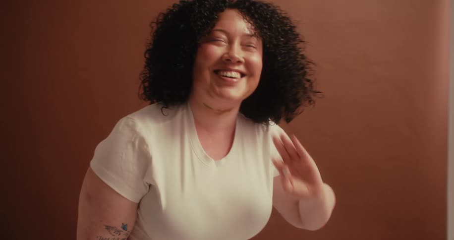 A chubby woman with curly hair wearing a white tshirt and a tattoo on her right arm poses and laughs joyfully