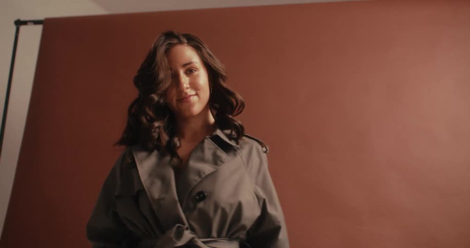 A woman with long flowing hair stands against a textured brown backdrop dressed in a gray trench coat
