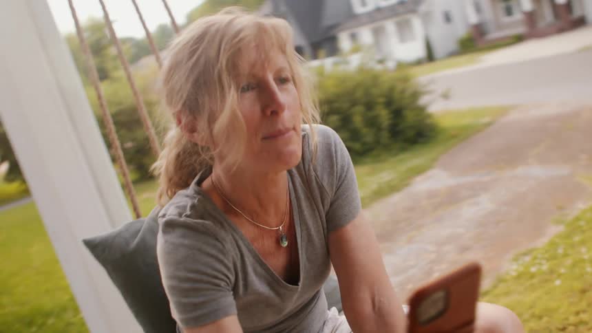 A woman with blonde hair in a ponytail sits on a porch swing holding a smartphone in her hand