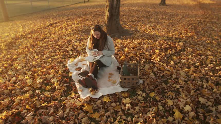 A young woman sits sipping a drink and reading under a tree in a park blanketed with fallen autumn leaves enjoying a peaceful moment