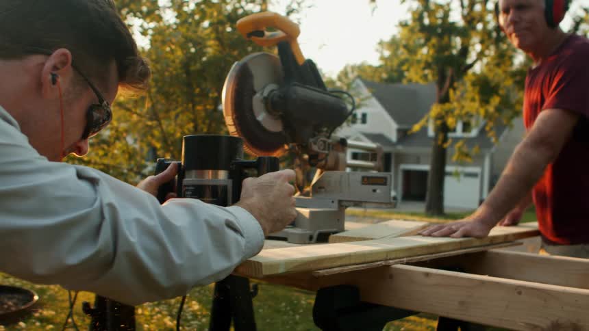 Two men are working outdoors at midday with one holding a saw to cut a piece of wood while the other secures the wood in place