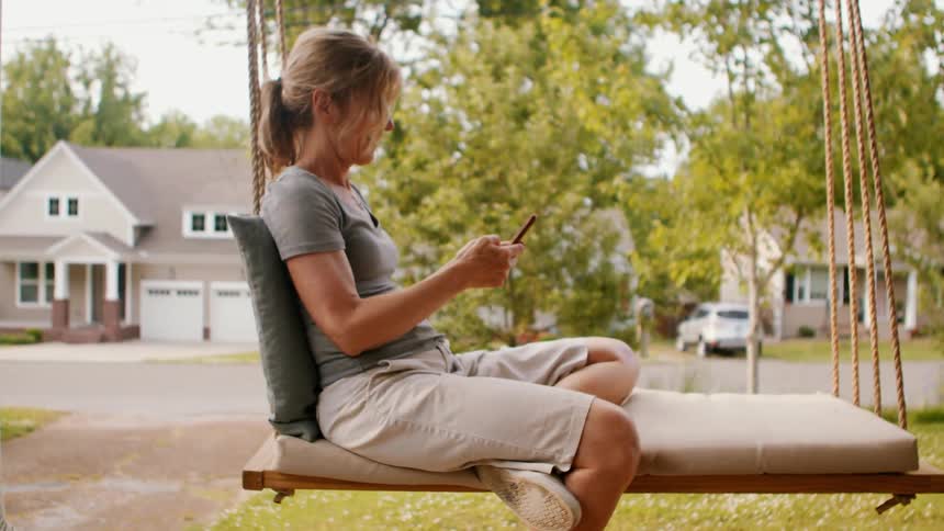 A woman looks at her smartphone while rocking on a front porch swing