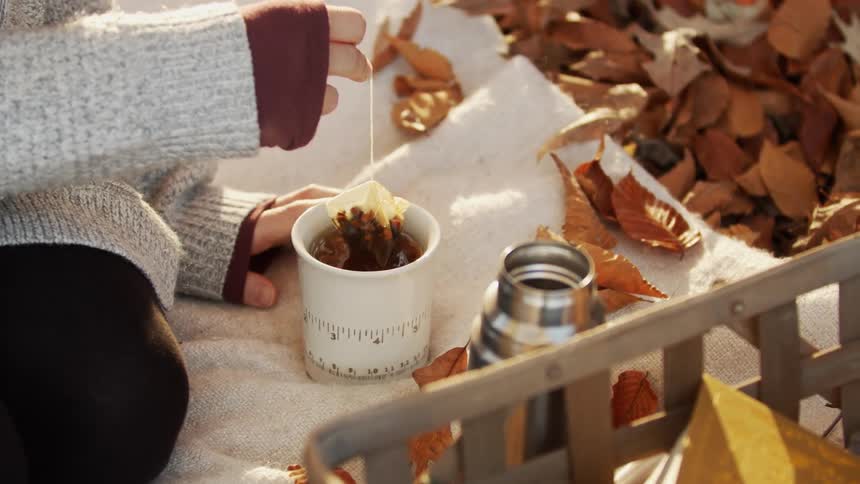 A woman is sitting on a blanket outside amongst the fallen leaves and making a cup of tea