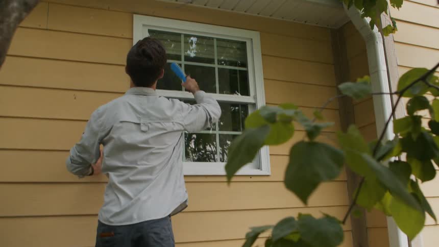 A woman tosses a man a brush and he cleans the siding of the house