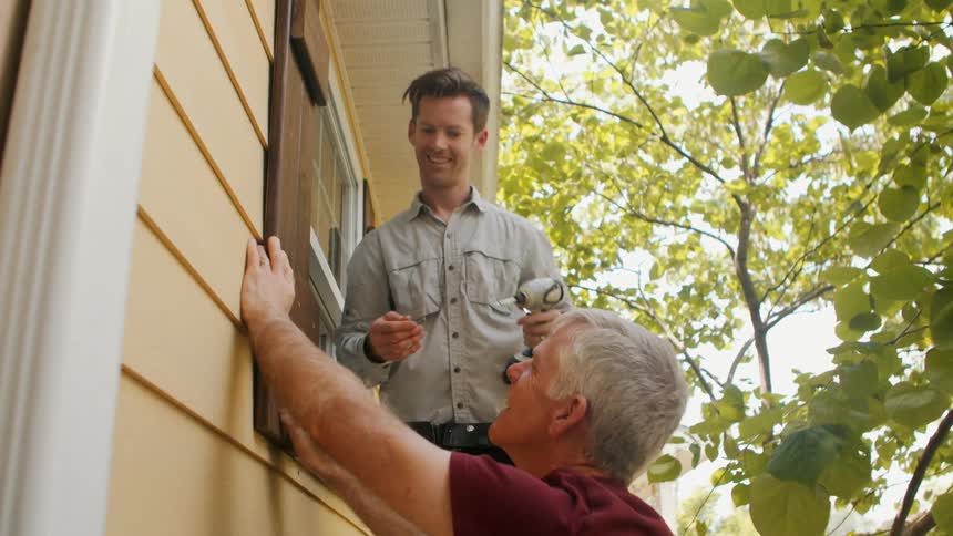 A man on a small ladder using a drill on a piece of wood by the window while another man holds the wood