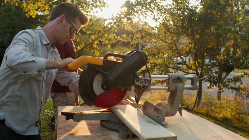 A man using a table saw outdoors to cut a piece of wood while an older man stands nearby watching