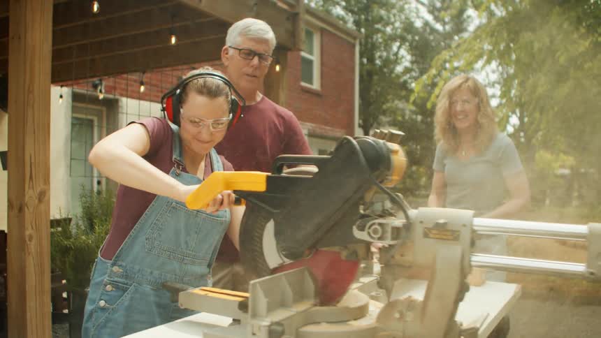 Woman uses a chainsaw under the supervision of a man while another woman watches smiling in the background